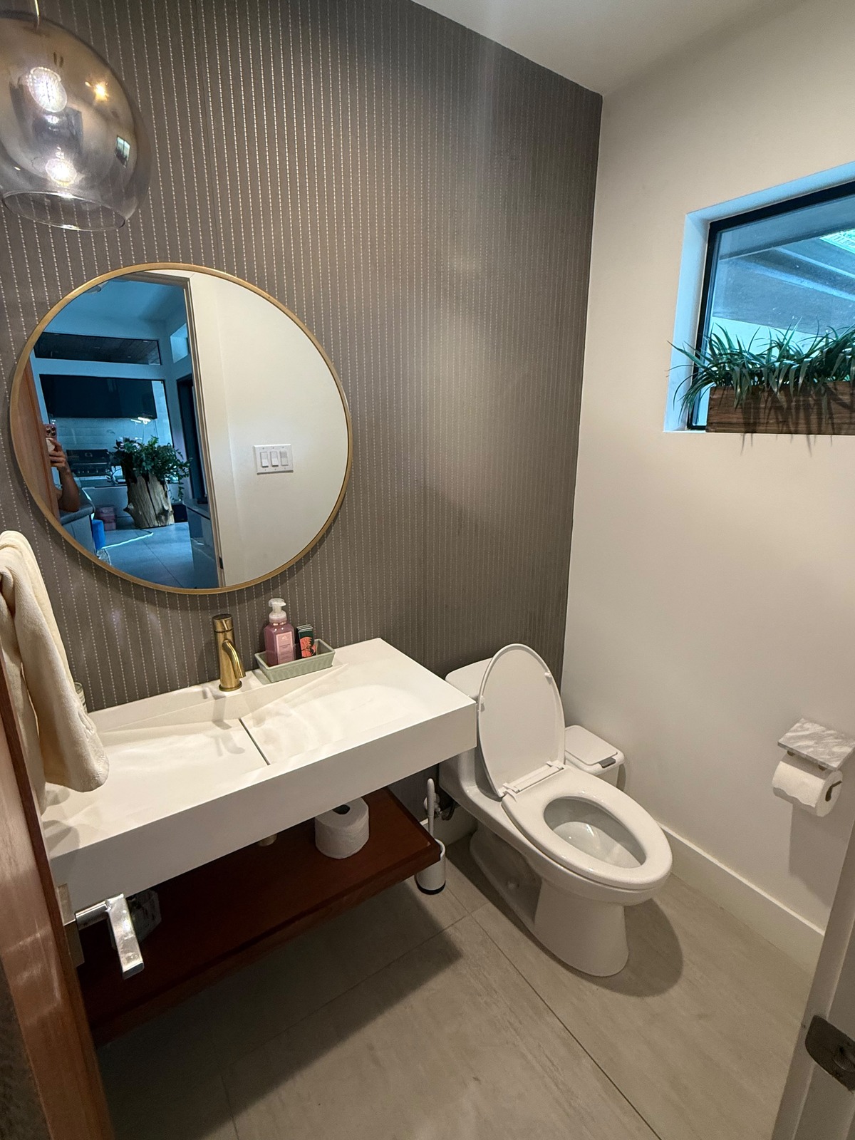 Powder bath with a taupe pinstripe wallpaper accent wall, white walls, a round gold mirror and a white floating sink